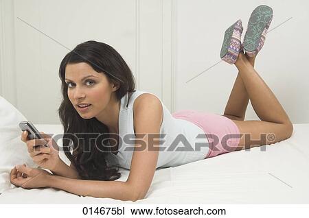 young woman lying on bed holding mobile phone and looking at camera View Large Photo Image Stock Image - young woman lying on bed holding mobile phone and looking at camera. Fotosearch