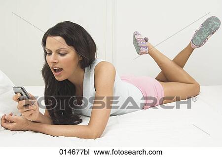 Stock Image - young woman lying on bed writing text messages in mobile. Fotosearch