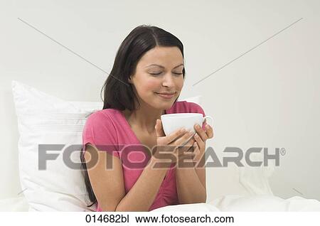 Stock Image - young woman sitting in bed smelling hot drink. Fotosearch