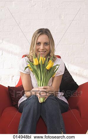 Stock Image - potrait of young woman sitting in armchair holding bunch of red tulips. Fotosearch