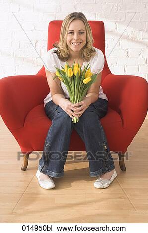 Stock Image - potrait of young woman sitting in armchair holding bunch of red tulips. Fotosearch