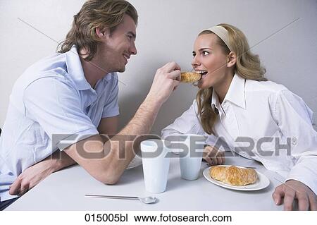 Stock Image - young couple having breakfast man feeding woman croissant. Fotosearch