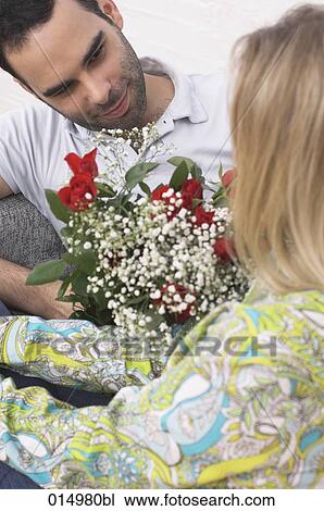 Stock Image - young couple sitting on armchair man giving red roses to woman. Fotosearch