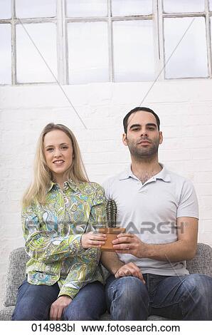 Stock Image - young couple sitting on armchair together holding potted cactus. Fotosearch