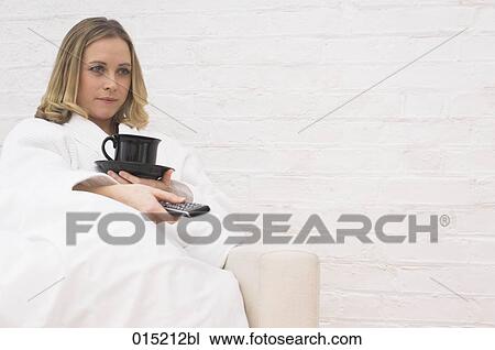 Stock Image - young woman in bathrobe sitting in arm chair holding cup and remote control. Fotosearch