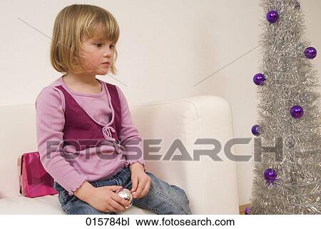 Stock Image - little girl sitting on sofa near christmas tree looking sad. Fotosearch