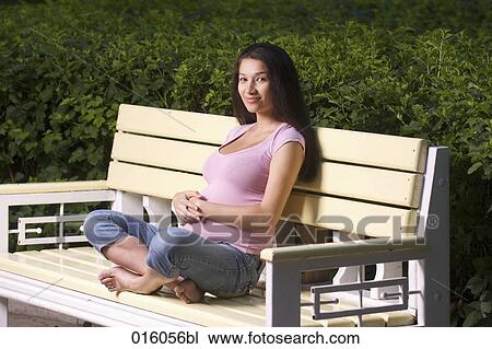 Stock Image - full body portrait of pregnant young woman sitting on park bench. Fotosearch