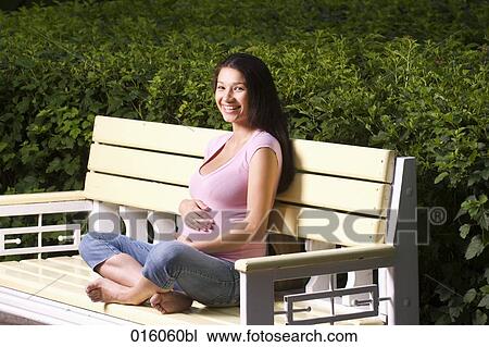 Stock Image - full body portrait of pregnant young woman sitting on park bench. Fotosearch