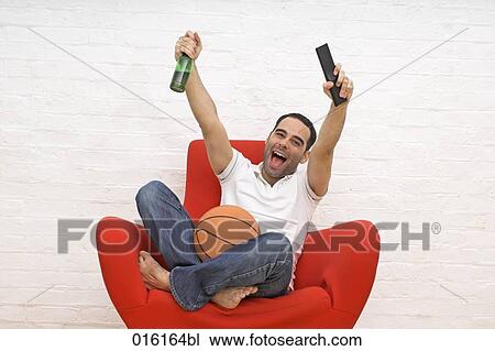 joyous young man sitting in red arm chair with basketball beer bottle and remote control watching tv View Large Photo Image Stock Image - joyous young man sitting in red arm chair with basketball beer bottle and remote control watching tv. Fotosearch