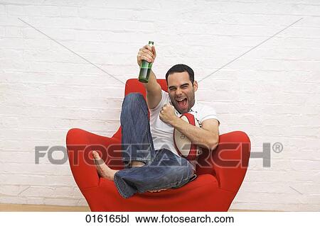 joyous young man sitting in red arm chair with rugby ball beer bottle and remote control watching tv View Large Photo Image Stock Image - joyous young man sitting in red arm chair with rugby ball beer bottle and remote control watching tv . Fotosearch