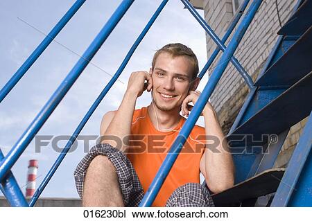 Stock Image - portrait of smiling young man sitting on stairs listening to music on mp3 player. Fotosearch