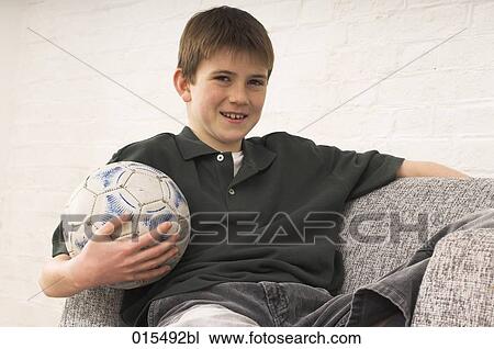 Stock Image - portrait of young boy sitting in arm chair holding football. Fotosearch