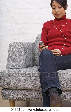Stock Image - portrait of young japanese woman sitting in arm chair listening to music on ipod. Fotosearch