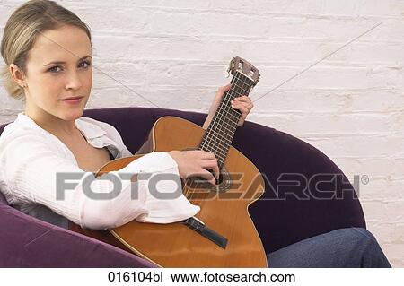Stock Image - portrait of young woman sitting at home playing guitar. Fotosearch