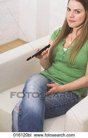 Stock Image - portrait of young woman sitting in arm chair holding remote control. Fotosearch