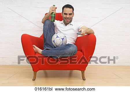 young man sitting in red arm chair holding football and beer bottle while watching tv View Large Photo Image Stock Image - young man sitting in red arm chair holding football and beer bottle while watching tv . Fotosearch