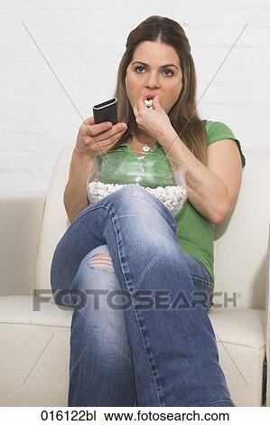 Stock Image - young woman watching tv holding remote control and eating popcorn. Fotosearch