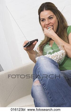 Stock Image - young woman watching tv holding remote control and eating popcorn. Fotosearch