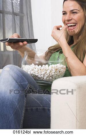 Stock Image - young woman watching tv holding remote control and eating popcorn. Fotosearch