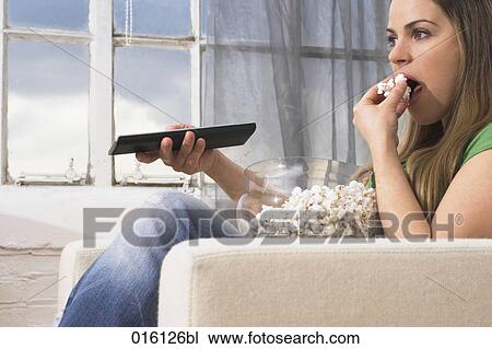 Stock Image - young woman watching tv holding remote control and eating popcorn. Fotosearch