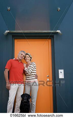 Stock Image - full body portrait of mature couple with dog standing at front door of their home. Fotosearch
