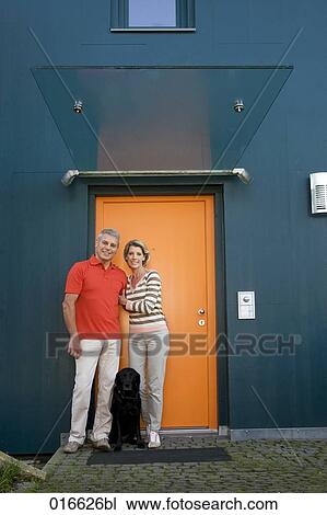 Stock Image - full body portrait of mature couple with dog standing at front door of their home. Fotosearch