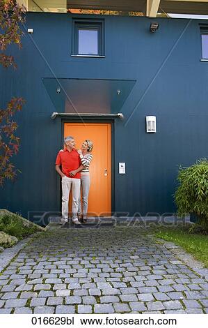 Stock Image - full body portrait of mature couple standing at front door of their home. Fotosearch