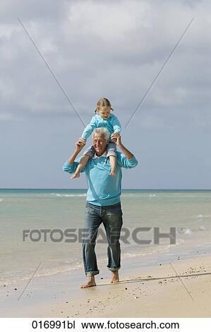Stock Image - grandfather carrying young girl piggyback on beach. Fotosearch