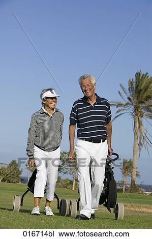 Stock Image - mature couple pulling golf bags over golf course. Fotosearch