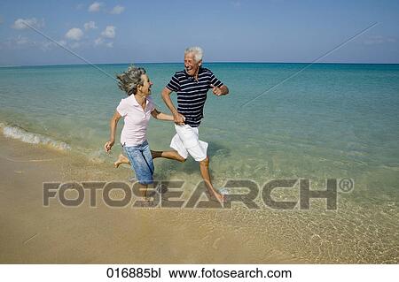 Stock Image - mature couple running along beach hand in hand. Fotosearch