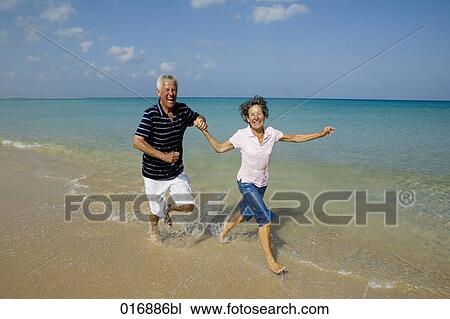 Stock Image - mature couple running along beach hand in hand. Fotosearch