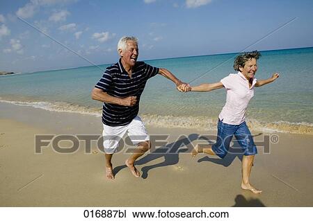 Stock Image - mature couple running along beach hand in hand. Fotosearch
