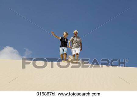 Stock Image - mature couple running down beach hand in hand. Fotosearch