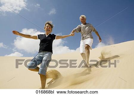 Stock Image - mature couple running down beach hand in hand. Fotosearch