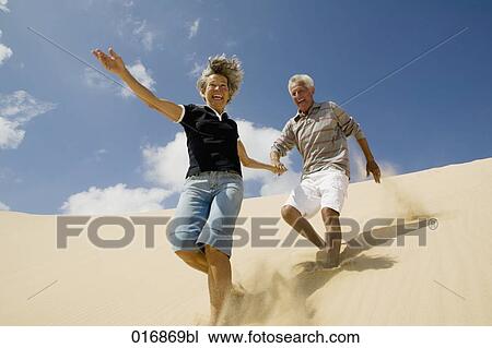 Stock Image - mature couple running down beach hand in hand. Fotosearch