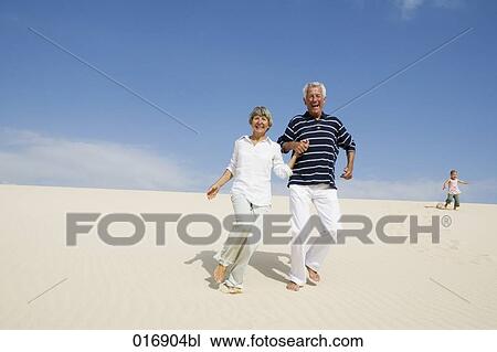Stock Image - mature couple running down beach hand in hand. Fotosearch