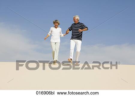 Stock Image - mature couple running down beach hand in hand. Fotosearch