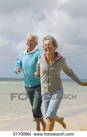 Stock Image - mature couple running hand in hand on beach. Fotosearch