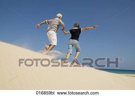 Stock Image - mature couple running on beach hand in hand. Fotosearch