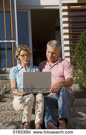 Stock Image - mature couple sitting in front of home with laptop computer. Fotosearch