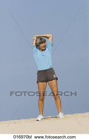 Stock Image - mature woman doing stretching exercises on sandy beach. Fotosearch