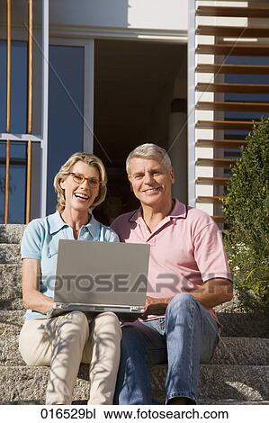 Stock Image - portrait of mature couple sitting in front of home with laptop computer. Fotosearch