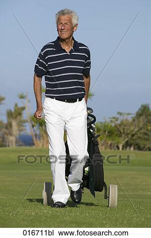 Stock Image - portrait of mature man pulling golf bag over golf course. Fotosearch