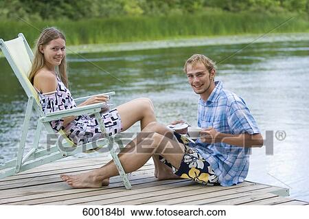 Stock Image - portrait of young couple sitting on pier drinking coffee. Fotosearch