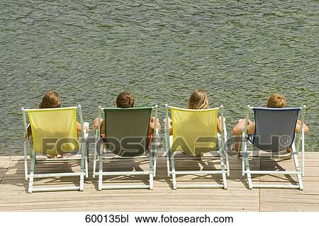 Stock Image - rear view of group of friends sitting in sunloungers on pier. Fotosearch