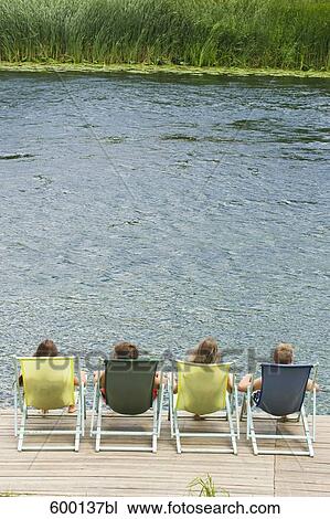 Stock Image - rear view of group of friends sitting on sunloungers on pier. Fotosearch