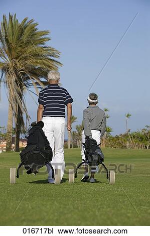 Stock Image - rear view of mature couple pulling golf bags over golf course. Fotosearch