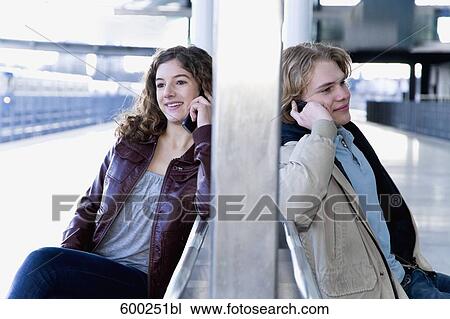 Stock Image - teenage couple sitting on bench at station talking on mobile phones. Fotosearch