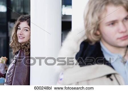 Stock Image - teenage girl leaning against pillar watching boy. Fotosearch