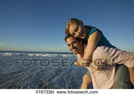 Stock Image - young man giving woman piggyback ride on beach. Fotosearch